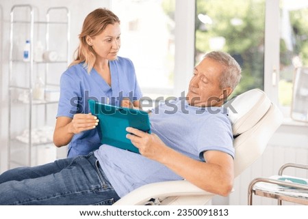 Old male patient lying on medical chair signing papers while talking with woman doctor