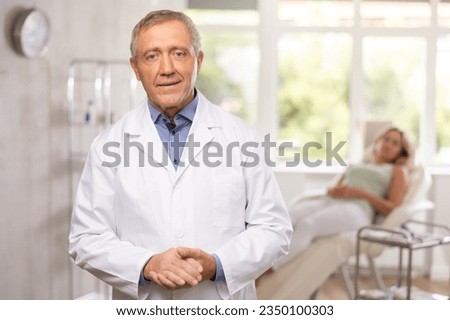 Positive old man doctor posing against background of doctor's cabinet with clinical chair Royalty-Free Stock Photo #2350100303