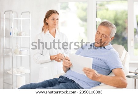 Female nurse shows clinic senior man client papers for familiarization and signing of consent for injection cosmetology procedure. Medical documentation, questionnaire, patient survey