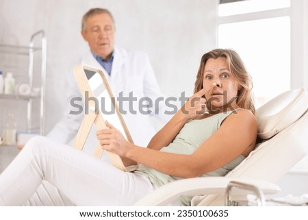 In cosmetology clinic, upset woman frowns, touches nose with finger, bad result of procedure, embarrassed doctor in background. Result did not meet expectations, unsatisfied client of plastic surgeon Royalty-Free Stock Photo #2350100635