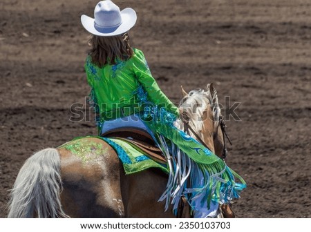 A cowgirl at a rodeo is wearing in green costume and sitting on the back of a blond horse. She has on a white hat. The horse has green glitter on its back. The arena is dirt.