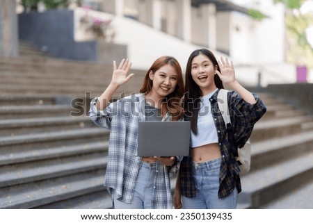 Two Asian female college students waving and looking forward and holding laptop computers on campus. Royalty-Free Stock Photo #2350139435