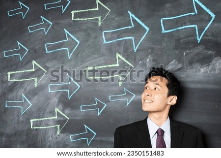 Happy Asian business man standing in front of a dark chalkboard with arrow signs drawn pointing.  Royalty-Free Stock Photo #2350151483