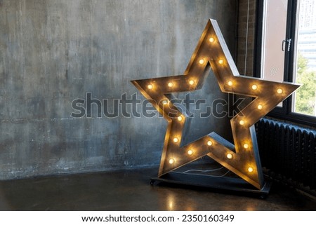 Illuminated star shaped wooden decoration stands in room by dark concrete wall and window. Copy space for your text or decoration. Soft focus. Photoshoot theme.