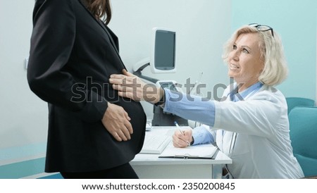 Confident doctor examines young pregnant woman in luxury clinic. Blonde therapist in white coat palpates belly of patient checking condition of foetus in office Royalty-Free Stock Photo #2350200845