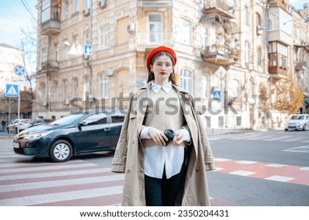 Happy young caucasian woman in good mood walks with coffee around city during day. Brunette wears glasses and red beret. Real emotions concept