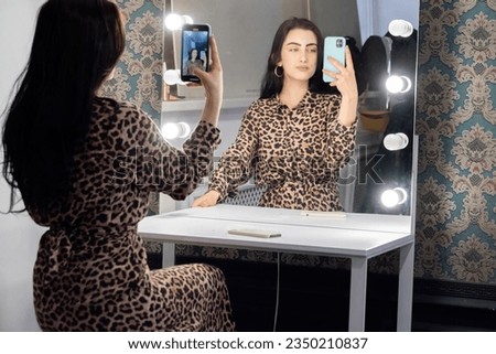 Woman in long leopard dress sitting at dressing table with phone in hand. Brunette tries to make selfie picture in mirror reflection