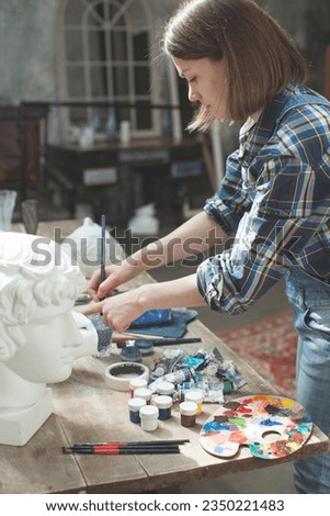 Interior of artist's workshop. Creative person concept. Easel. Woman paints a picture. The artist chooses colors for the painting. She is wearing denim overalls and plaid shirt. Mock up.