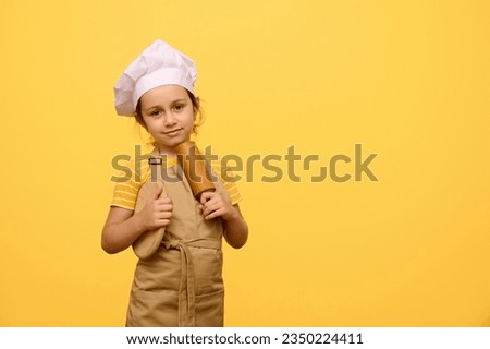 Studio portrait of adorable charming Caucasian little baker confectioner girl in beige apron and hat, holding a wooden board and rolling pin, smiles cutely looking at camera, isolated yellow backdrop
