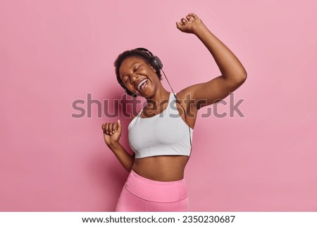 Joyful African woman loses herself in rhythm of her music moves gracefully with her eyes closed shakes arms and laughs happily wears tracksuit isolated over pink background. Dance to your own melody