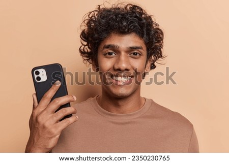 Photo of handsome curly haired young Hindu man holding modern phone playing video game surfing internet being in good mood dressed casually isolated over brown background. Technology concept