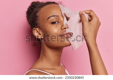 Beautiful woman with curly combed hair puts on facial mask for skin treatment smiles gently wears t shirt isolated over pink wall. Young female model removing from face skincare hydrating sheet mask Royalty-Free Stock Photo #2350230845