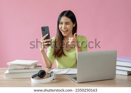 A positive and pretty Asian girl shows her thumb up and enjoys talking on video call with her friend while sitting at her study table against an isolated pink background.