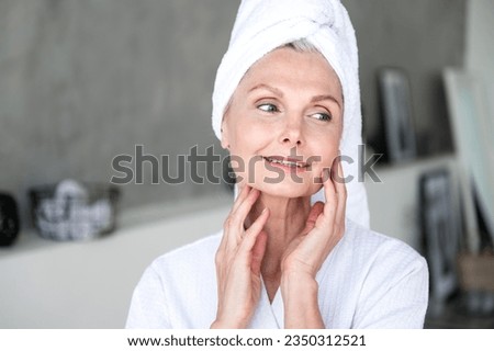 50 years old smiling woman with towel on head looking away, touching moisturized soft healthy sensitive skin on cheek and neck, doing morning hydration, spa beauty treatment. Face care concept Royalty-Free Stock Photo #2350312521