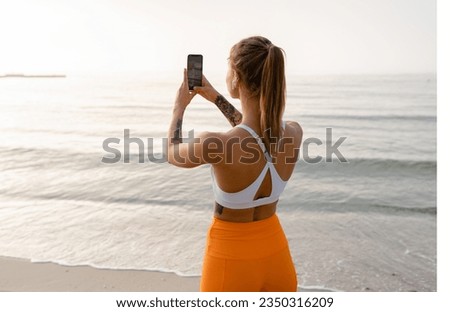pretty young smiling woman doing sports in the morning by the sea beach in stylish sport outfit sportswear, healthy fit lifestyle, using smart phone taking photo on camera