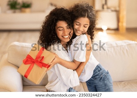 Mother's day concept. Adorable latin girl giving gift and embracing mom, mommy and daughter bonding together at home, sitting on sofa