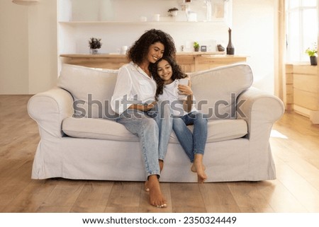 Adorable latin mother and daughter using cellphone, sitting on sofa in living room, spending time together at home, having video chat, copy space