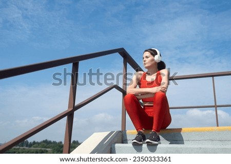 female athlete in sportswear wearing wireless headphones and listening to music, sitting on steps resting. Outdoor fitness concept.