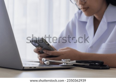 Young female doctor with eye glasses using mobile smartphone and work on laptop computer with medical stethoscope on the office desk in room at clinic or hospital.