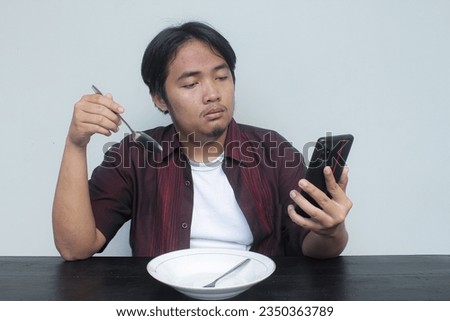 Young Asian man in red shirt sitting in front of empty plate and looking at cellphone and showing bored and sad expression. Eat concept.