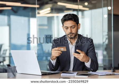 Successful young arab businessman inside office at workplace, man serious thinking and concentrated making online money transfer, boss holding bank credit card in hands, using phone app.