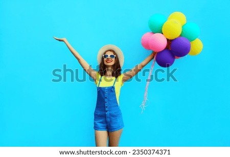 Summer image of happy laughing young woman with bunch of colorful balloons having fun wearing straw hat on blue studio background