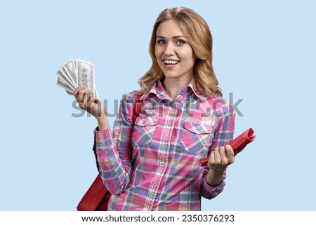 Portrait of blonde woman holding fan of dollars. Red purse and wallet. Isolated on pastel blue background.