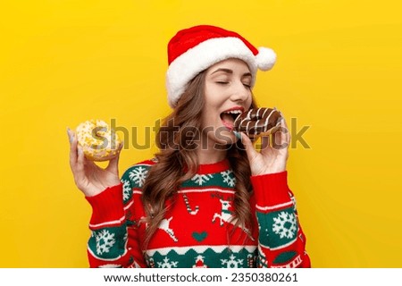 girl in Christmas sweater and santa claus hat holds two donuts and eats on yellow isolated background, woman with sweet pastries in New Year's clothes