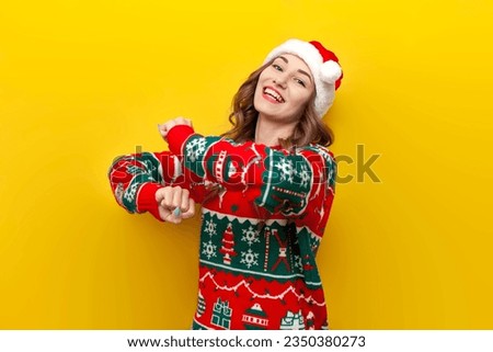 young girl in Christmas sweater and santa claus hat smiles and dances on yellow isolated background, woman in New Year's clothes moves her hands to the music in disco