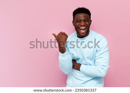 african american man in blue sweater laughs and points his hand to the side on pink isolated background, man makes fun and jokes and advertises copy space