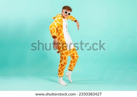 young funny guy in orange giraffe pajamas dances at party in sunglasses on blue isolated background, man in animal costume moves to music, pajama disco concept