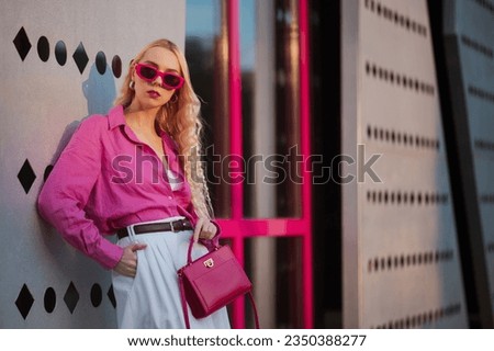 Fashionable confident blonde woman wearing trendy pink sunglasses, linen shirt, white bermuda shorts, holding small classic faux leather bag, posing in street of city. Copy, empty space for text Royalty-Free Stock Photo #2350388277