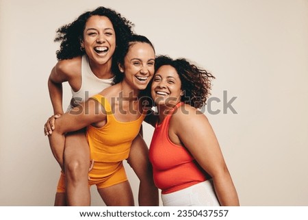 Happy female friends celebrate a healthy lifestyle through sports, exercise and fitness. Fit young women standing together in a studio, dressed in fitness attire and smiling at the camera. Royalty-Free Stock Photo #2350437557