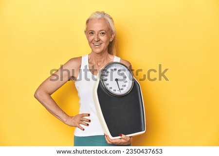 Sporty middle-aged Caucasian woman holding scale, yellow studio laughing and having fun.