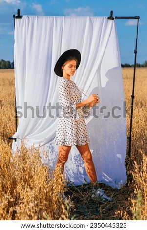 Rural Countryside Scene. Young beautiful woman with long hair dressed in white Polka-dot dress and black hat standing at golden oat field with a white curtain on the background.