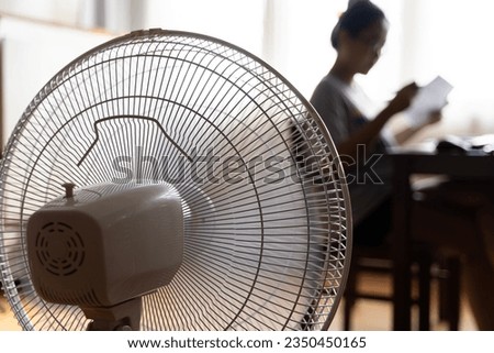 Sunny summer in front of the working fan, suffering from the summer heat. Unhappy Asian woman sitting in front of fan at work suffering from heat in modern house. Royalty-Free Stock Photo #2350450165