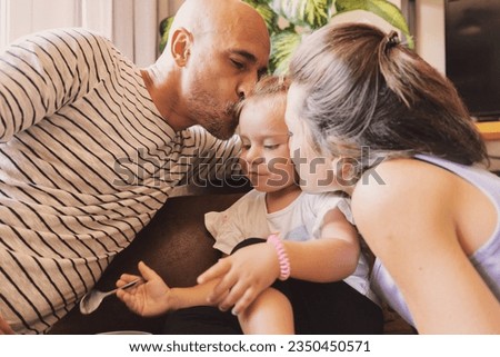 Dad and mom smooch their baby girl while she eats baby food with spoon, loving family on sofa in living room