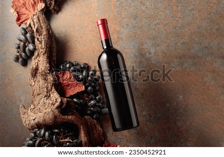 Bottle of red wine in motion on a rusty background with an old snag, blue grapes, and dried-up vine leaves. Concept of vinery. Copy space.