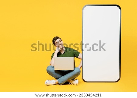 Full body young IT man wears green t-shirt casual clothes big huge blank screen mobile cell phone smartphone with workspace area hold use work on laptop pc computer isolated on plain yellow background