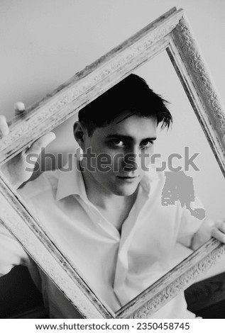 The black-white photograph captures a young handsome man in a white shirt sitting next to a white wall, holding an empty picture frame in his hands. The connection between the artist and his craft.