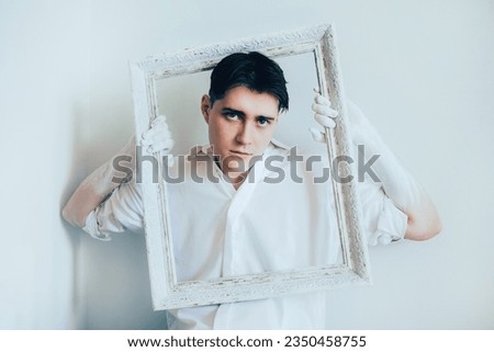 A handsome young man wearing a white shirt, holding a blank white frame for a painting. The role of the artist in creating something beautiful out of nothingness. Framing in art.