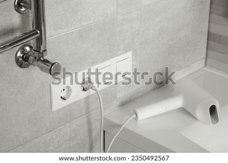 Hairdryer plugged into power socket on light grey wall in bathroom Royalty-Free Stock Photo #2350492567