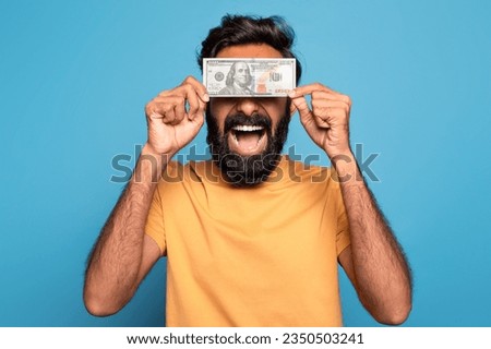 Easy money. Excited indian man holding dollar banknote and cover eyes, posing on blue studio background. Wealth and income, financial success, cash greediness concept Royalty-Free Stock Photo #2350503241