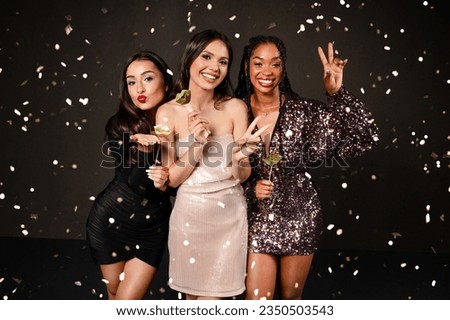 Cheerful attractive multicultural ladies in nice outfits posing together among falling confetti over black background, holding photo booth pops, grimacing and gesturing. New Year party