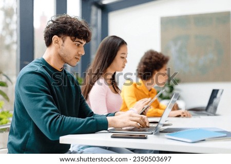Multiracial male and female students studying for test, making homework with gadgets, focused guy using laptop, sitting at table in coworking space