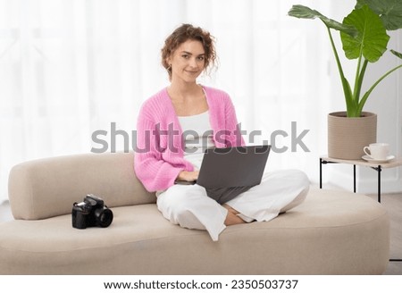 Portrait Of Smiling Young Woman With Laptop Sitting On Couch Next To Photocamera, Beautiful Millennial Freelance Photographer Lady Using Computer At Home For Editing Photos, Copy Space