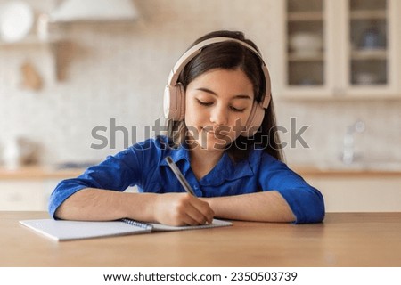Arabic schoolgirl learning and doing homework, writing or drawing in her paper notebook and wearing wireless headphones sitting at table in home interior. Kid girl taking notes studying in kitchen