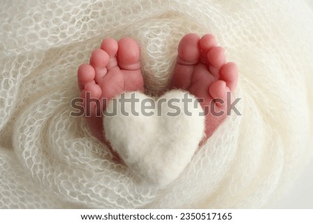 Knitted white heart in the legs of a baby. The tiny foot of a newborn baby. Soft feet of a new born in a white wool blanket. Close up of toes, heels and feet of a newborn. Macro photography