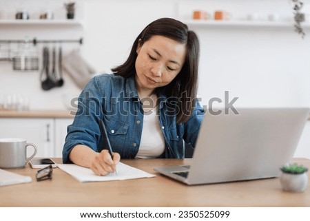 Close up view of asian woman making notes while reading article on portable computer in spacious room. Focused female adult in casual wear using online educational resources for writing exam test. Royalty-Free Stock Photo #2350525099