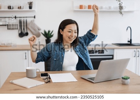 Thrilled asian woman raising hands in winner's gesture while looking at digital screen of computer in apartment. Emotional worker rejoicing over excellent news related to business partnerships. Royalty-Free Stock Photo #2350525105
